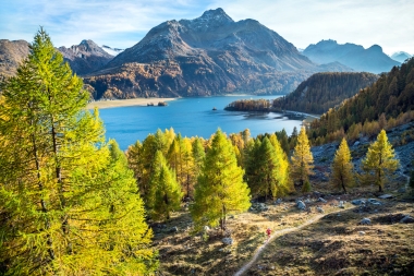 A picturesque mountain landscape with a lake, surrounded by autumn-colored trees and a hiker on a trail.