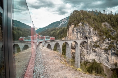 A red train is traveling over a viaduct through a mountainous landscape.