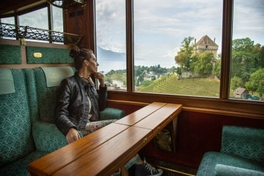 Eine Frau sitzt in einem Zugabteil und blickt nachdenklich aus dem Fenster auf eine Landschaft mit Weinbergen und einem Schloss.