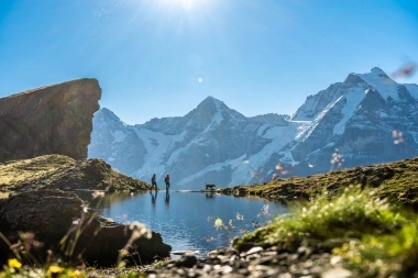Eine Berglandschaft mit einer Wasserstelle, zwei Wanderern und einem Hund im Vordergrund.