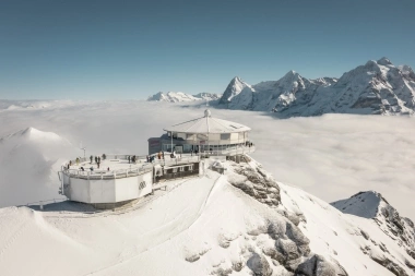 Eine runde Bergstation auf einem verschneiten Gipfel, umgeben von Wolken und schneebedeckten Alpen.