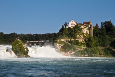 A castle on a hill above a large waterfall, with a train on a bridge in the background.