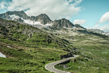 Eine kurvige Bergstraße vor einer Gebirgskulisse mit schneebedeckten Gipfeln und grünem Gras.