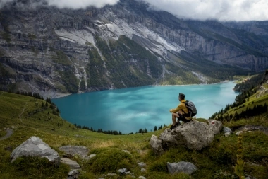 Una persona seduta su una roccia guarda un lago di montagna turchese, circondato da montagne e cielo nuvoloso.