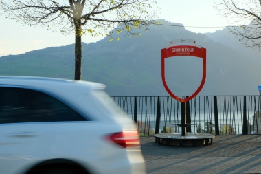Eine weisse Limousine fährt an einem 'Grand Tour of Switzerland' Schild vorbei, im Hintergrund Berge und ein See.