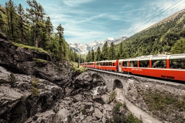 Ein roter Zug fährt über eine Steinbrücke in einer alpinen Landschaft mit Bergen und Bäumen.