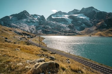 Binari ferroviari si snodano lungo un lago limpido, circondati da montagne innevate sotto un cielo blu.