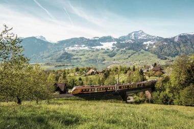 Un train jaune-orange traverse un paysage vert et vallonné avec des montagnes enneigées à l'arrière-plan.