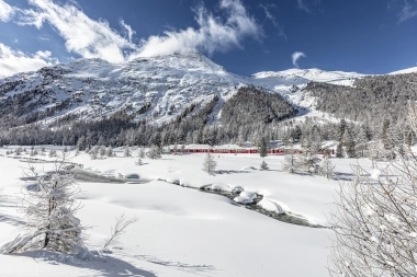 Eine rote Bahn fährt durch eine verschneite Berglandschaft mit klarem Himmel.