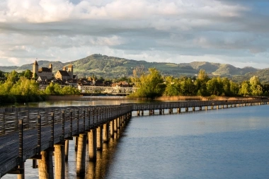 Eine lange Holzbrücke führt über einen ruhigen See, im Hintergrund eine malerische Stadt und grüne Hügel.