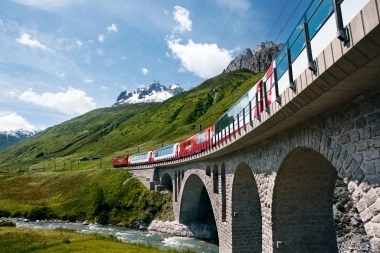 A red train travels on a viaduct through a green mountain landscape with snow-capped peaks.