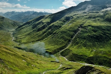 Un paesaggio montano verde con un piccolo fiume e una locomotiva fumante.