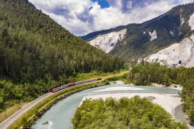 Ein roter Zug fährt durch eine grüne, bergige Landschaft entlang eines Flusses.