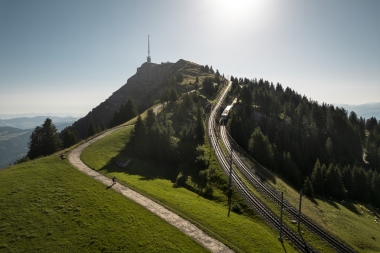 Eine Berglandschaft mit einer Eisenbahnstrecke, die zu einem Gipfel mit einem Sendemast führt, umgeben von Bäumen und Wiesen.