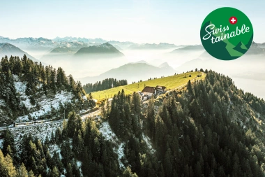 Panoramic view of the Swiss Alps with forests, mountains, and a small farmhouse, overlaid with a 'Swisstainable' logo.