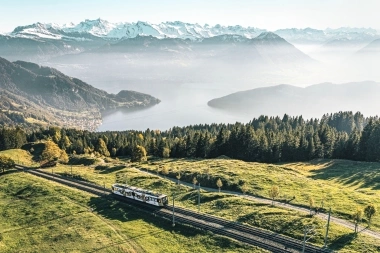 Un train traverse des collines vertes avec des montagnes et un lac en arrière-plan.