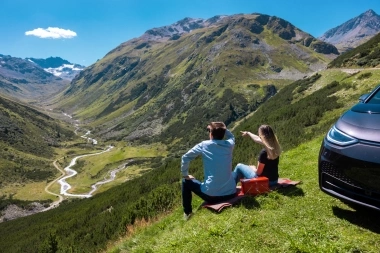 Two people sit on a meadow overlooking a valley in the mountains, next to a parked car.