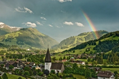 Eine Kirche in einem grünen Tal mit Bergen im Hintergrund und einem Regenbogen.