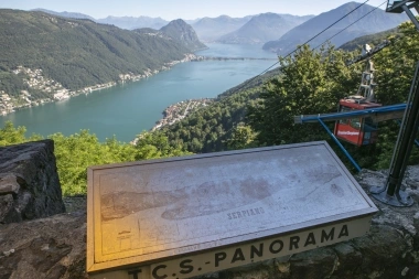 Eine Panoramatafel auf einem Aussichtspunkt zeigt einen See und umliegende Berge; eine Seilbahn ist sichtbar.