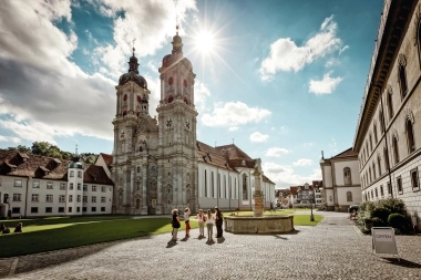 A Baroque cathedral with two towers, surrounded by old buildings and a square with fountains; people are walking in sunny weather.