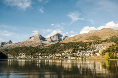 Eine ruhige Berglandschaft mit einem See im Vordergrund und einem Dorf an den Hängen unter klarem, blauem Himmel.