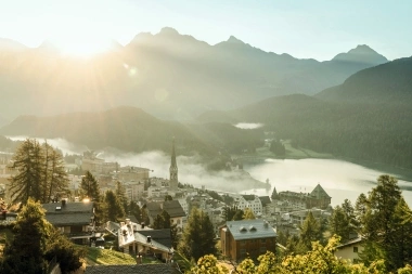 Tramonto su una cittadina alpina con chiesa, circondata da montagne e un lago.