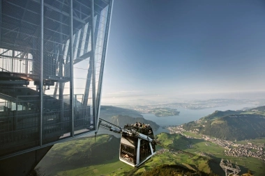 A cable car ascends the mountain overlooking a lake and a city in the valley.