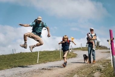 Une famille marche sur un chemin, tandis qu'un homme et un enfant sautent joyeusement ; une femme porte un petit enfant.