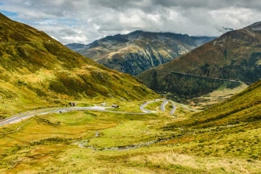 Eine kurvige Bergstrasse windet sich durch grüne Hügel und Täler unter einem bewölkten Himmel.