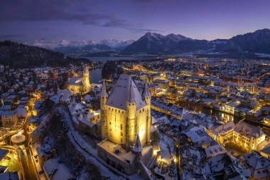 A winter city at night, illuminated buildings, snow-covered roofs, and mountains in the background.
