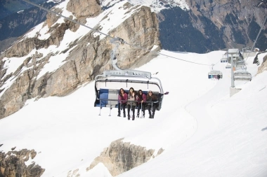 A group of people sitting on a chairlift in a snowy mountain landscape.