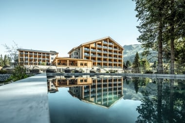 A modern hotel building is reflected in a calm pool, surrounded by mountains and trees.