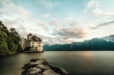 An old castle on the lakeshore, surrounded by mountains under a cloudy sky.