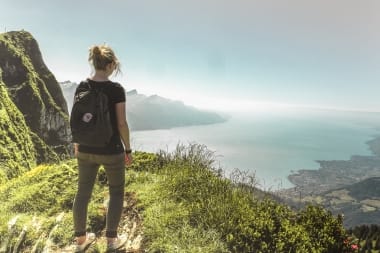 A woman with a backpack stands on a mountain and looks at a lake and surrounding landscape.