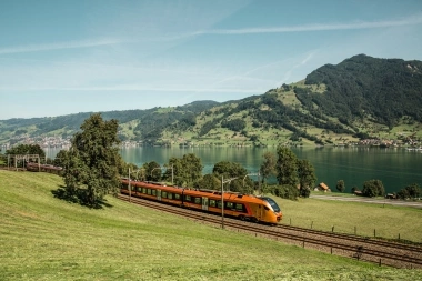 An orange train travels along a lake, surrounded by green hills.