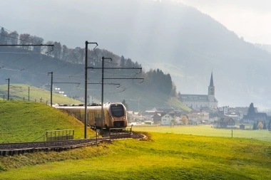Treno su un binario ferroviario attraverso un paesaggio verde con un villaggio sullo sfondo.