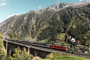 A red locomotive on a viaduct in front of a mountain landscape.
