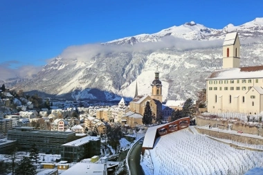 Un paesaggio urbano invernale con edifici innevati e montagne sullo sfondo.