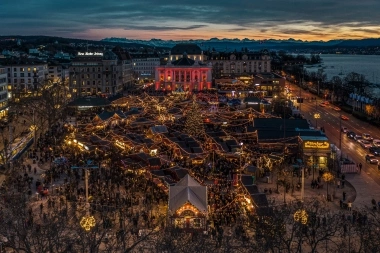 Aerial view of a festively lit Christmas market at dusk, surrounded by buildings with a view of mountains and a lake.