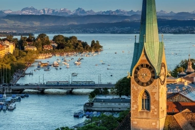 A church with a clock tower, view of the lake with boats and mountains in the background.