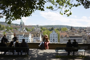 Eine Gruppe von Menschen sitzt auf Bänken mit Blick auf eine Stadtlandschaft bei sonnigem Wetter.