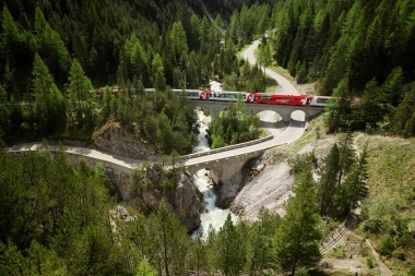 Eine rote Bahn überquert eine Brücke in einer bewaldeten Berglandschaft mit einem Fluss darunter.