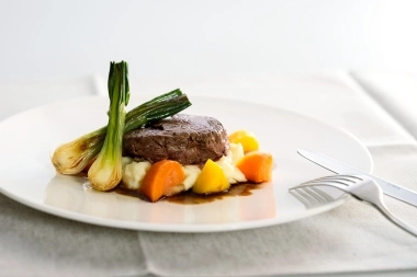 Ein Teller mit Rinderfilet, Frühlingszwiebeln, Karotten und Kartoffelpüree auf einer weissen Serviette.