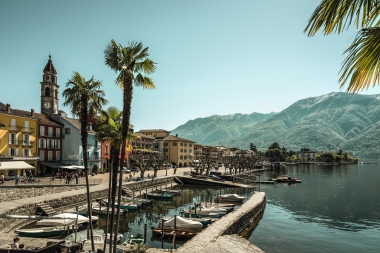 A beautiful harbor promenade with palm trees, colorful buildings, and boats on the water; mountains in the background.