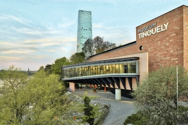 Museum Tinguely with modern architecture, surrounded by trees, with a tall glass tower in the background.