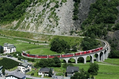 Un treno rosso sta attraversando un ponte di pietra curvo in un paesaggio montano verde.