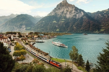 Paysage de montagne avec lac, train et bateau au premier plan.