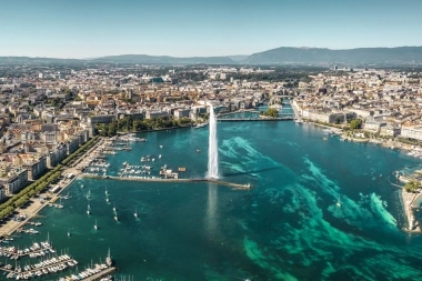 View of Geneva with the Jet d'Eau fountain in the foreground and Lake Geneva, surrounded by buildings and mountains in the background.