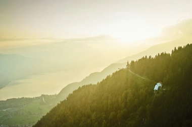 View of a wooded hill with a chapel at sunset, with a lake and valley in the background.