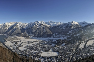 Weite Berglandschaft mit schneebedeckten Gipfeln und einem Tal mit Ortschaften, unter blauem Himmel.